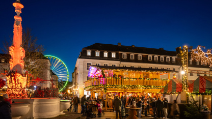 Christkindl-Markt auf dem St. Johanner Markt Der St. Johanner Markt ist weihnachtlich geschmückt mit Lichtern und Ständen. Links ist der Marktbrunnen festlich beleuchtet, im Hintergrund erscheint hinter den Häusern die Hälfte des neonfarben leuchtenden Riesenrads auf dem Tbilisser Platz.